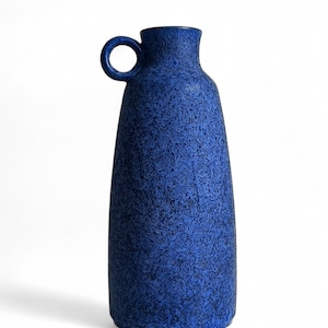 Puede incluir: Un jarrón azul oscuro texturizado con un asa de bucle. El jarrón tiene un cuello estrecho y una base más ancha. El color azul tiene un efecto moteado. El asa es un pequeño bucle circular. El jarrón es probablemente de cerámica o gres.