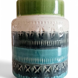 May include: A ceramic jar with a green, white, blue and black geometric pattern. The jar has a wide mouth and a rounded body.