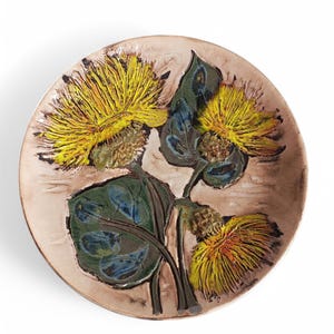 Puede incluir: Un plato de cerámica con un diseño en relieve de tres flores amarillas con hojas verdes. El plato tiene un fondo marrón claro.
