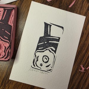 May include: A black and white linocut print of a stylized object with a rectangular shape and a circular design. The print is on white paper, with the artist's signature, "Melissa Ulin", in the lower left corner. A pink linoleum block is visible.