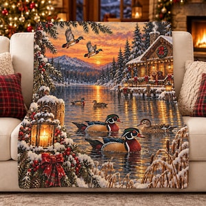 May include: A rectangular throw blanket featuring a winter landscape with a snow-covered cabin, a lake, and several ducks. The scene includes a glowing lantern in the foreground, evergreen branches with red berries, and a sunset sky.