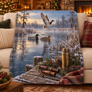 Peut inclure: Un plaid doux illustrant une scène hivernale avec trois canards colverts sur un lac enneigé. L'image montre un ponton en bois avec un thermos en métal, deux appeaux en bois, des pommes de pin et un bord en tissu écossais sur un paysage gelé.