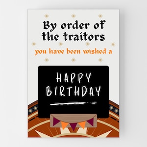 May include: A birthday card with a cream background and text that reads "By order of the traitors you have been wished a Happy Birthday." The card has a black chalkboard with white text and orange and purple pennants.