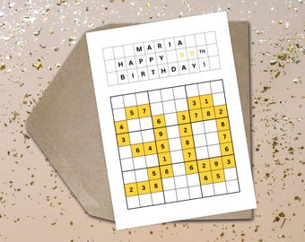 May include: A white birthday card with a Sudoku puzzle and the text "MARIA HAPPY BIRTHDAY! 90th". The card is on a brown envelope with gold confetti.