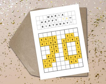 May include: A white birthday card with a Sudoku puzzle and the text "MARIA HAPPY BIRTHDAY! 70 TH". The Sudoku grid has yellow highlighted numbers. The card is on a brown envelope with gold confetti.