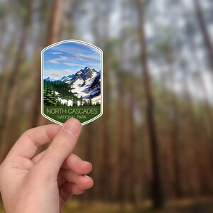 May include: A sticker featuring a scenic view of a mountain range with snow-capped peaks and a blue sky with clouds. The sticker is green and white with the text "NORTH CASCADES NATIONAL PARK" in black.