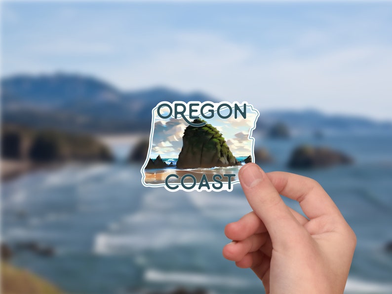 May include: A die-cut sticker featuring a scenic illustration of a large sea stack rock formation on a sandy beach. The text OREGON COAST is printed in dark blue capital letters at the top and bottom of the sticker against a coastal landscape.