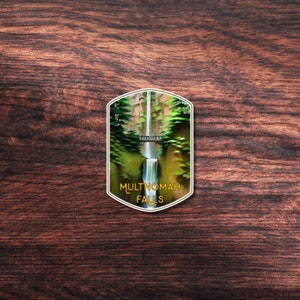 May include: A vintage-style illustrated patch with a green and brown image of Multnomah Falls in Oregon. The patch has a gold border and the text "Multnomah Falls" in gold lettering.