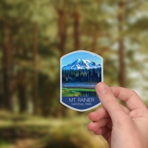 May include: A blue and white sticker with a picture of Mount Rainier National Park. The sticker features a mountain range with snow-capped peaks and a lake in the foreground. The text "MT. RAINIER NATIONAL PARK" is printed in white on a blue background.