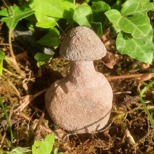 May include: A small, light brown, carved stone sculpture shaped like a mushroom with a rounded cap and a thick, cylindrical stem. The object has a textured, granular surface and is positioned upright, displaying a solid, uniform earthy color.