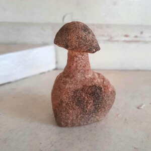May include: A small, hand-carved stone figurine shaped like a mushroom with a rounded cap and a thick, sturdy stem. The object features a rough, porous texture with a natural reddish-brown color and darker, mottled patches across its surface.