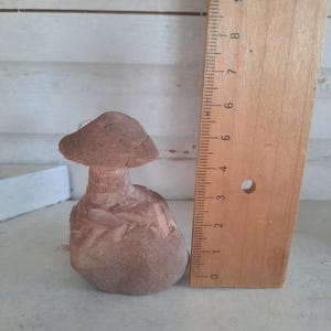 May include: A brown, carved stone mushroom figurine with a textured cap and stem, positioned next to a wooden ruler. The ruler indicates the mushroom stands approximately 4.5 inches tall, showing clear markings in inches along its vertical edge.