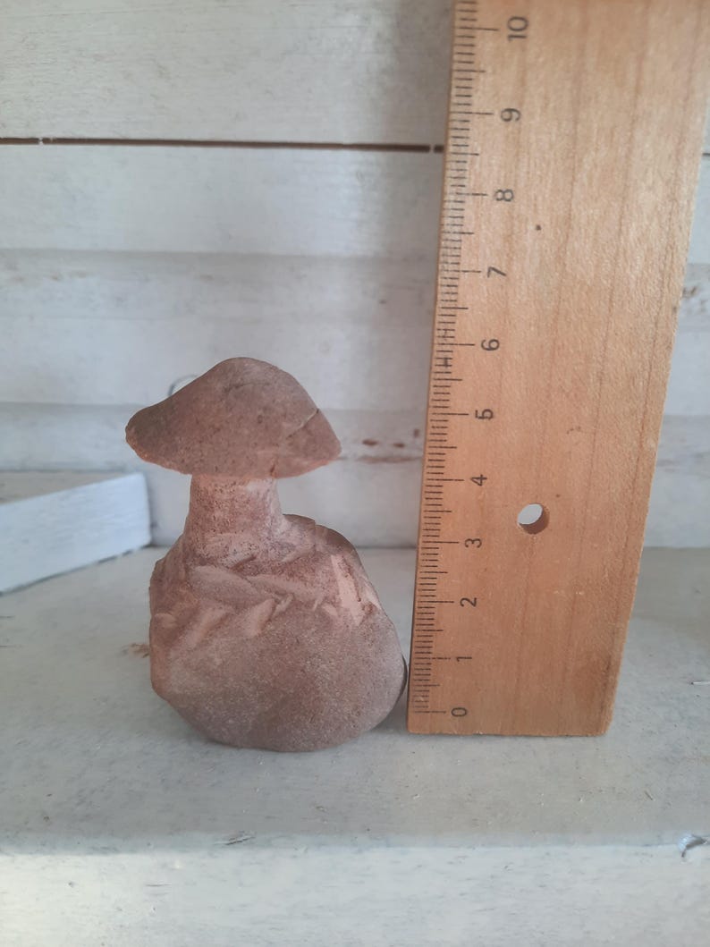 May include: A brown, carved stone mushroom figurine with a textured cap and stem, positioned next to a wooden ruler. The ruler indicates the mushroom stands approximately 4.5 inches tall, showing clear markings in inches along its vertical edge.