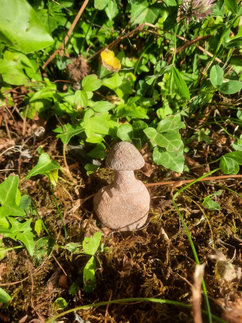 May include: A small, light brown, carved stone sculpture shaped like a mushroom with a rounded cap and a thick, cylindrical stem. The object has a textured, granular surface and is positioned upright, displaying a solid, uniform earthy color.