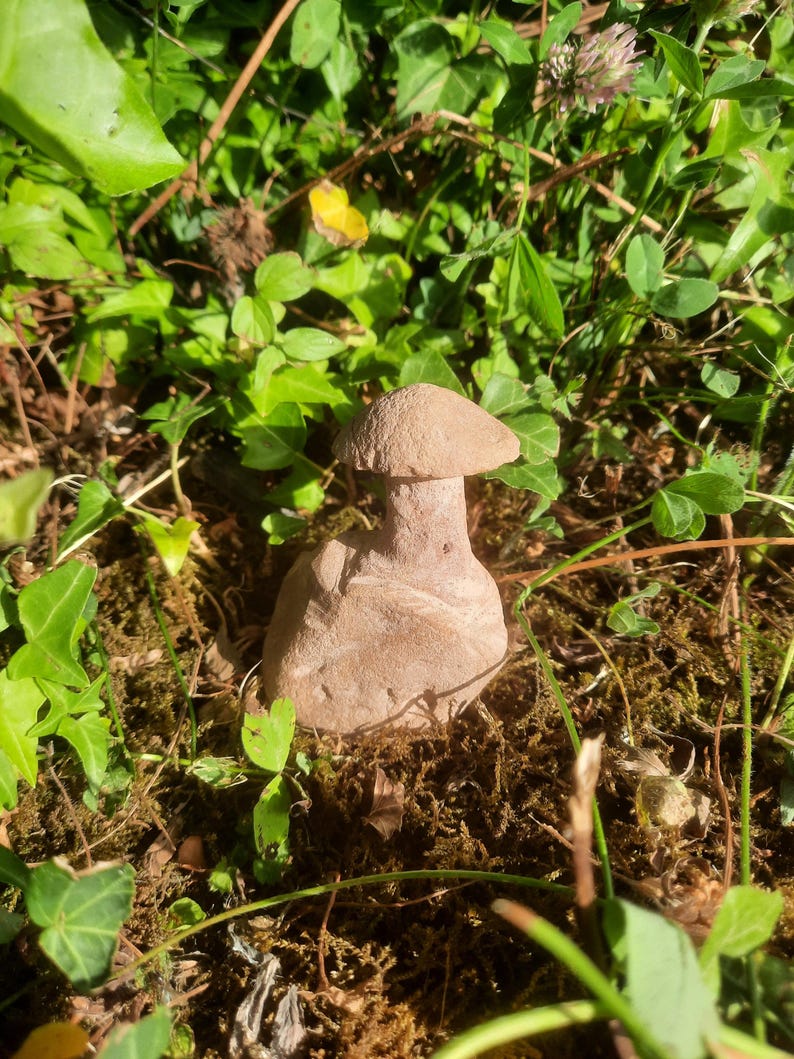 May include: A small, light brown, carved stone sculpture shaped like a mushroom with a rounded cap and a thick, cylindrical stem. The sculpture has a smooth, matte texture and sits upright, displaying natural variations in the stone's color and grain.