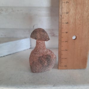 May include: A small, textured, reddish-brown stone figurine shaped like a mushroom. The object is positioned next to a wooden ruler, which indicates that the figurine stands at a height of approximately 3.5 inches from base to cap.