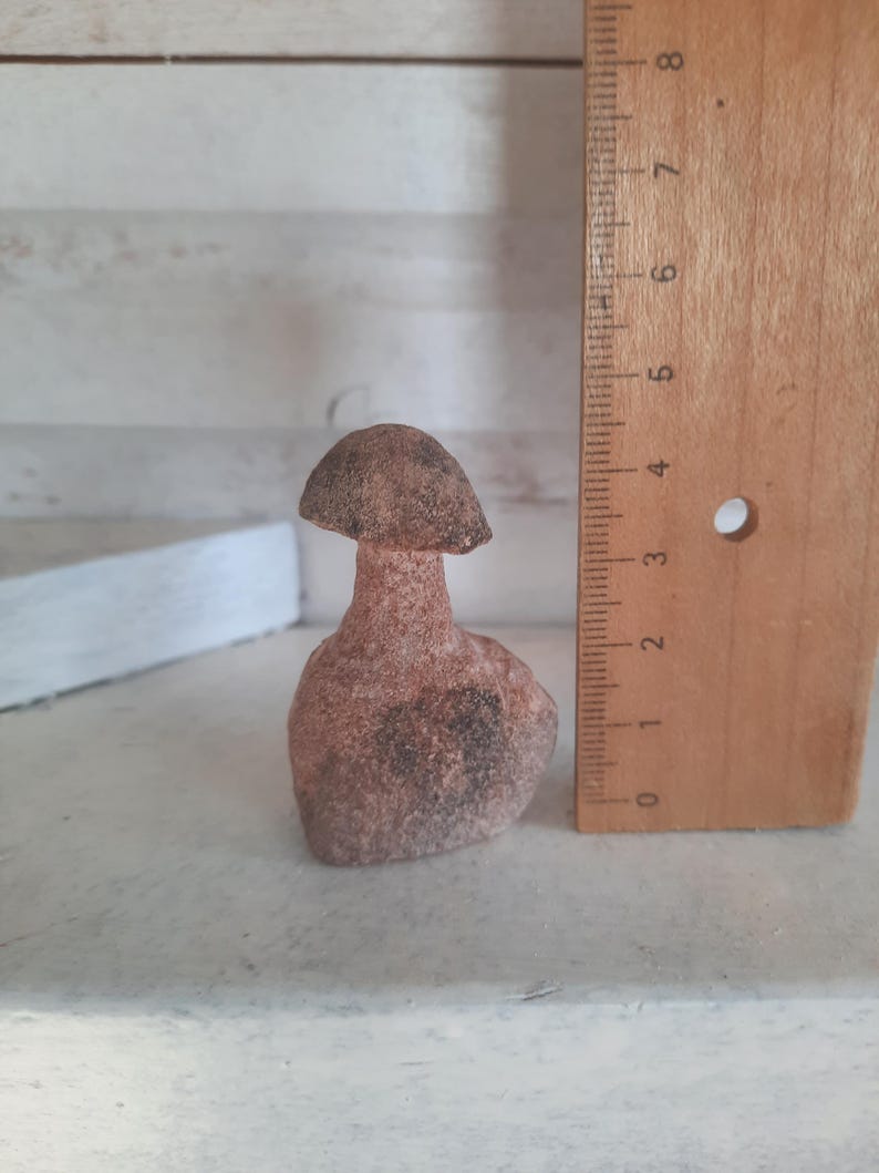 May include: A small, textured, reddish-brown stone figurine shaped like a mushroom. The object is positioned next to a wooden ruler, which indicates that the figurine stands at a height of approximately 3.5 inches from base to cap.