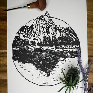 May include: Black and white linocut print of a mountain landscape within a circle. The print depicts a mountain range, trees, and a body of water. A wood carving tool and sprigs of lavender and a thistle are arranged around the print.