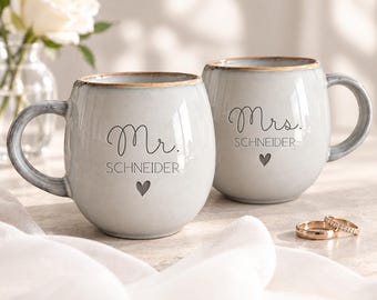 May include: Two light gray ceramic mugs with a rustic brown rim. One mug features the text Mr. SCHNEIDER with a small heart icon, and the other features Mrs. SCHNEIDER with a small heart icon. Two gold-toned wedding bands sit nearby.