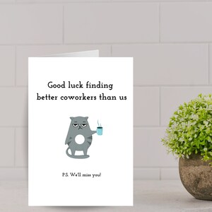 May include: A white card with a gray cat cartoon holding a blue cup of coffee. The text on the card reads "Good luck finding better coworkers than us." and "P.S. We'll miss you!"