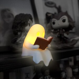 May include: A small, white, glowing ghost figurine sitting on a shelf edge while reading a brown book. The ghost has a smooth, rounded head and emits a warm yellow light from its eyes and body, contrasting with the surrounding dark shelf.