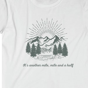 May include: A white short-sleeve t-shirt featuring a dark gray graphic of a mountain range, pine trees, and a sunburst. Below the illustration, the text reads: It's another mile, mile and a half. The design is centered on the chest of the shirt.