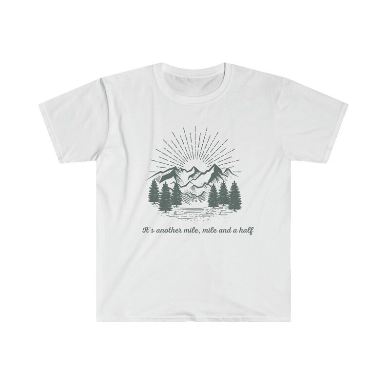 May include: A white short-sleeve t-shirt featuring a dark gray graphic of a mountain range, pine trees, and a sunburst. Below the illustration, the text reads: It's another mile, mile and a half. The design is centered on the chest of the shirt.