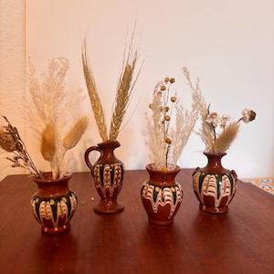 Può includere: Quattro piccoli vasi decorativi in ceramica con fiori secchi e spighe di grano. I vasi sono marroni con motivi verdi, neri e bianchi. Sono disposti su un tavolo di legno scuro su sfondo neutro.