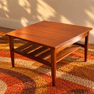 May include: A square wooden coffee table with a smooth, medium-brown finish. The table features four tapered legs and a lower slatted shelf for storage. The surface is flat and rectangular, showcasing a natural wood grain pattern throughout the piece.