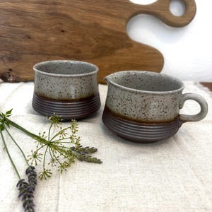 Puede incluir: Dos tazas de cerámica con una sección superior gris claro moteada y una base inferior acanalada de color marrón oscuro. Una taza tiene una pequeña asa y un pico vertedor, mientras que la otra es redonda. Acabado esmaltado liso.