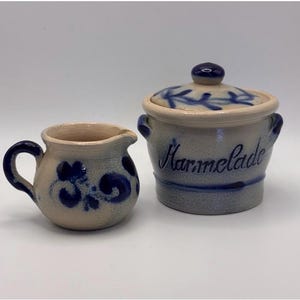 Op de afbeelding: Een kleine keramische melkkan en een pot met deksel met het woord "Marmelade" in blauw schrift. Beide stukken zijn beige met blauwe bloemmotieven. De pot heeft een kleine knop op het deksel en twee handvatten.