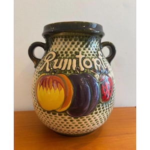 May include: A ceramic vase with a textured, dotted pattern. The vase features a dark green rim and handles. It is decorated with colorful illustrations of a yellow pumpkin, a purple eggplant, and a red strawberry. The word "Rumtopf" is written across the top.