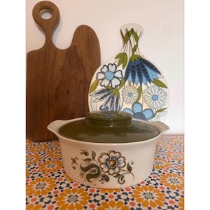 Può includere: Una pirofila vintage bianca e verde a motivi floreali con coperchio abbinato. Un tagliere in legno e un tagliere floreale decorativo sono sullo sfondo. La pirofila ha una base color crema con un motivo floreale.