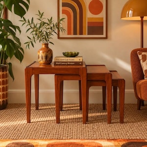 May include: A set of three nested wooden tables with a smooth finish. The top table holds a brown ceramic vase, a small green glass bowl, and two books titled The Kinfolk Home and Mid-Century Modern. The tables feature tapered legs.