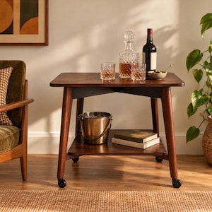 May include: A dark wood rolling bar cart with a rectangular top and a lower shelf. The top holds a crystal decanter, two matching glasses, a wine bottle, and a small bowl. The lower shelf contains a metallic ice bucket and two stacked books.