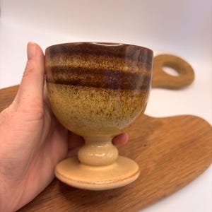 May include: A handmade ceramic goblet with a pedestal base. The cup has a brown and tan glaze with horizontal bands. The goblet is held in a hand, with a wooden cutting board in the background.