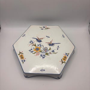 May include: A white, hexagonal ceramic serving platter with a blue border. The platter features a hand-painted design of two blue birds, yellow and blue flowers, and small insects. The platter is supported by small feet.