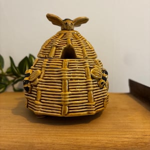 May include: A ceramic honey pot shaped like a beehive, with a lid and a bee-shaped handle. The pot is tan with brown accents and has two bee figures on the sides. The beehive is sitting on a wooden surface.