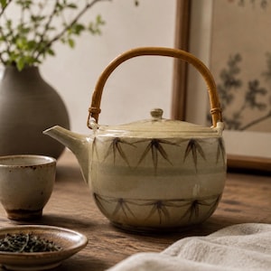 May include: A ceramic teapot with a light beige glaze and dark brown painted bamboo leaf motifs. The teapot features a curved spout, a matching lid, and a rounded handle made of light brown woven material attached to two small loops.