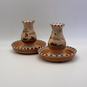 Könnte beinhalten: Zwei Kerzenhalter aus Keramik mit beigefarbener, gesprenkelter Oberfläche. Jeder Halter hat einen runden Sockel und eine vasenartige Oberseite, die mit einem Segelboot und einer Landschaft in Braun, Orange und Beige verziert ist. Die Ränder sind mit einem dunkelbraunen Muster verziert.