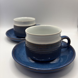 May include: Two ceramic coffee cups and saucers. The cups have a white rim and a blue body with horizontal brown stripes. The saucers are solid blue. The set is on a white background.