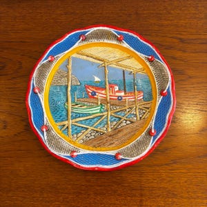 May include: A round ceramic decorative plate featuring a relief scene of a wooden dock with a red boat and a green boat on blue water. The border is blue with a textured net pattern, accented by a red rim and small red spheres attached by white cord.