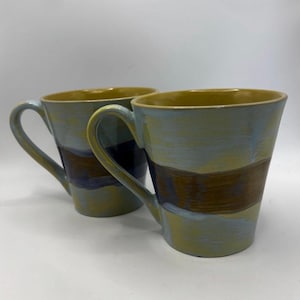 Könnte beinhalten: Zwei Keramikbecher mit handgemaltem Design. Die Becher haben ein hellblaues und braunes horizontales Streifenmuster. Der Innenraum und der Griff sind senfgelb. Die Becher sind für heiße oder kalte Getränke geeignet.