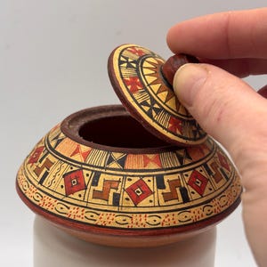 May include: A small, round, lidded ceramic jar with a hand-painted geometric design in red, black, and tan. The lid is being lifted to reveal the interior. The jar is resting on a white cylindrical base.