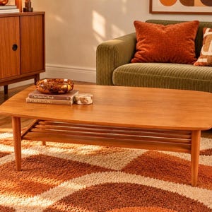 May include: A rectangular wooden coffee table with rounded corners and four tapered legs. The table features a slatted lower shelf and a smooth top surface holding a brown glass bowl and two stacked books titled The Interior Book and Period Living.