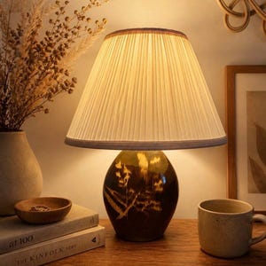 May include: A table lamp featuring a pleated lampshade and a dark brown base with a botanical design. The lamp is placed on a wooden surface, alongside a ceramic mug and a small wooden bowl. The lampshade is a light cream colour.