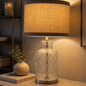 May include: A table lamp with a clear glass base featuring a textured, repeating scalloped pattern and a metallic silver base. The lamp is topped with a cylindrical fabric lampshade in a light tan colour with dark grey trim at the top and bottom edges.