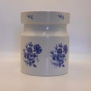 Könnte beinhalten: Ein weißer Keramiktopf mit blauen Blumenmustern. Der Topf hat eine zylindrische Form mit einem etwas breiteren oberen und unteren Teil. Blaue Blumenmuster sind symmetrisch auf dem Körper des Topfes angeordnet, mit kleineren floralen Akzenten am Rand.