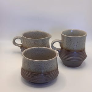 May include: A set of three handmade ceramic items: a mug, a small bowl, and a creamer. The pieces are speckled beige with a brown, ribbed base. The mug has a curved handle.