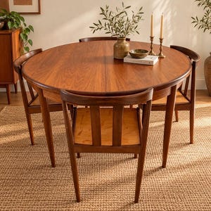 May include: A round wooden dining table with a visible grain pattern, accompanied by four matching wooden chairs with curved backrests. On the table sits a ceramic vase with green branches, a small bowl, and a book titled Kinfolk.